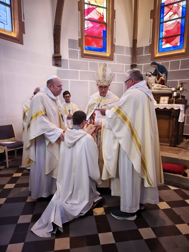 Colin kneels before Archbishop Karl-Michael Soemer during his ordination, accompanied by clergy