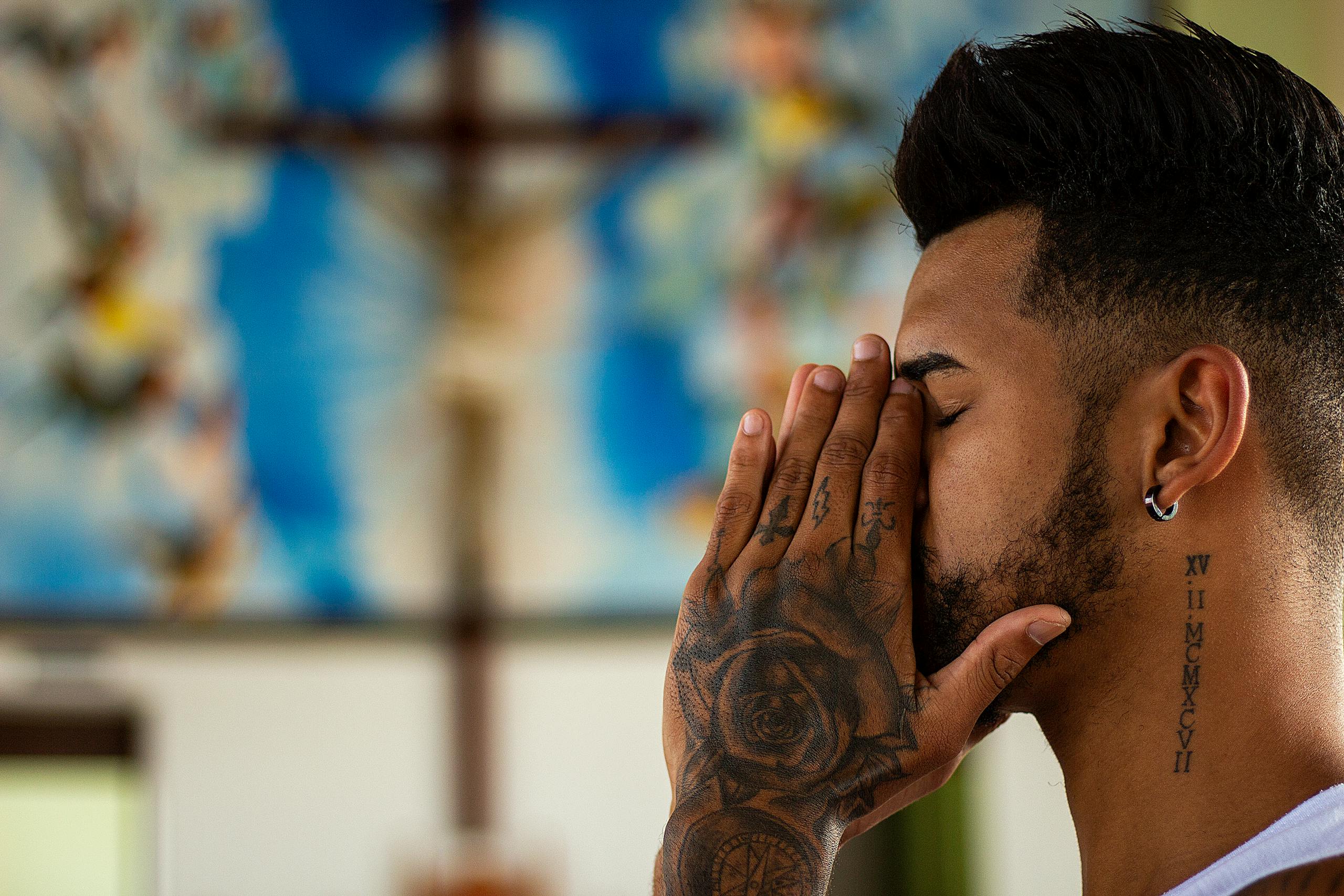 Apostolic Old Catholic Mission | Christ At The Centre. Everyone At The Table. 2 A man with tattoos prays inside a church, capturing a moment of reflection and spirituality.